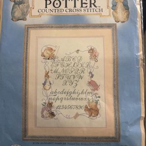May include: A counted cross stitch kit titled The World of Beatrix Potter featuring an alphabet sampler design. The packaging displays illustrations of Peter Rabbit and friends, with text indicating a finished size of 16.5 inches by 13.5 inches.
