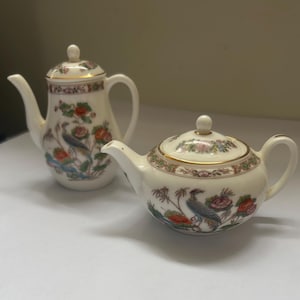 Könnte beinhalten: Eine cremefarbene Kaffeekanne aus Porzellan und eine passende Teekanne, beide mit einem zarten Blumenmuster mit Vogelmotiv, goldfarbenen Rändern und Deckeln mit kleinen kugelförmigen Knöpfen. Die Stücke zeigen detaillierte botanische und vogelartige Illustrationen.