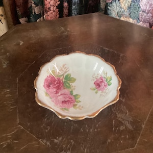 May include: A small, white porcelain bowl with a scalloped edge and a gold trim. The bowl is decorated with a floral pattern of pink roses and green leaves.