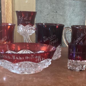 May include: Five vintage red glass cups and a bowl with etched designs and dates. The cups are all different sizes and shapes, and the bowl is round with a scalloped edge. The cups and bowl are all made of clear glass with a red tint.