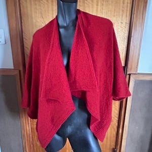 Könnte beinhalten: Ein leuchtend roter Schal-Wrap auf einer schwarzen Schaufensterpuppe. Der Wrap hat eine offene Front mit breiten Revers und kurzen Ärmeln. Der Stoff scheint eine strukturierte Wolle zu sein, die einen gemütlichen und stilvollen Look bietet. Ideal für kühle Tage.