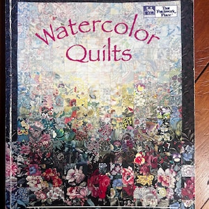 May include: A softcover book titled Watercolor Quilts by Pat Maixner Magaret and Donna Ingram Slusser. The cover features a grid of floral fabric squares transitioning from light to dark colors, framed by a dark paisley border.