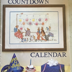 May include: A Beatrix Potter Countdown calendar featuring a cross-stitch illustration of animals at a birthday party with numbered candy attachments. Below, a blue party hat, a cupcake with a blue candle, and a blue gift box with a white bow.