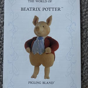 May include: A white paper print featuring The World of Beatrix Potter and Pigling Bland. The image shows a knitted pig figure wearing a red jacket, a blue and white striped vest, and a blue tie. The design includes light blue decorative scrollwork.
