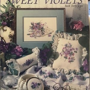 Può includere: Una collezione di oggetti a punto croce con il tema delle viole. L'immagine mostra un punto croce incorniciato, un cuscino, un contenitore rotondo e tessuto con ricami di viole. Il testo "Paula Vaughan's Sweet Violets" è visibile.