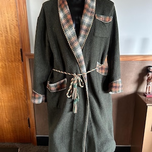 May include: A dark green dressing gown with a plaid collar, cuffs, and pocket details. It features a matching belt and tassel. The garment is displayed on a black mannequin.