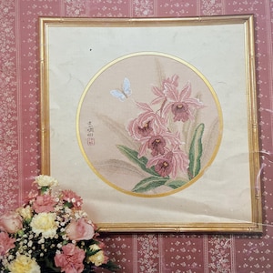 Può includere: Opera d'arte incorniciata intitolata "Ming Orchids" con un disegno circolare di orchidee rosa e una farfalla. L'opera d'arte con cornice dorata è ambientata su una carta da parati floreale rosa. Un mazzo di fiori è di fronte all'opera d'arte.