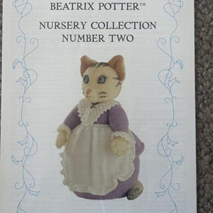 Puede incluir: Un folleto de papel blanco titulado BEATRIX POTTER NURSERY COLLECTION NUMBER TWO con una muñeca de gato tejida llamada MRS TABITHA TWITCHIT. El gato lleva un vestido morado con cuello de encaje blanco y delantal, enmarcado por un borde azul.