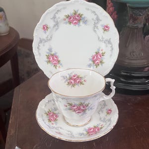 Peut inclure: Un ensemble de tasse et soucoupe en porcelaine blanche avec un motif floral de roses roses et de fleurs bleues. L'ensemble a un bord délicat et festonné et une bordure dorée.