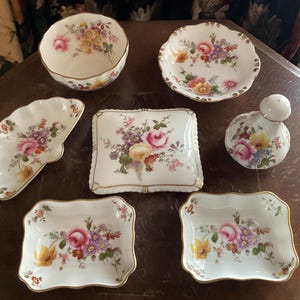 Puede incluir: Un juego de siete platos de porcelana vintage con motivos florales. Los platos son de diversas formas y tamaños, incluyendo un tazón redondo, un plato cuadrado y un plato en forma de media luna. Los platos están decorados con delicados diseños florales en tonos de rosa, amarillo y verde.