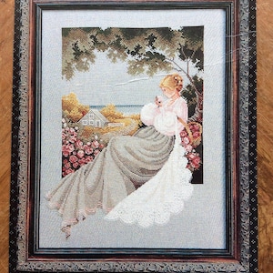 May include: Cross-stitch artwork of a woman in a long dress seated outdoors, framed in a decorative wooden frame. The scene includes a house, flowers, and a tree, with a soft colour palette.