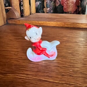 May include: A ceramic figurine of an elf wearing a red hat and red clothing, sitting on a white and pink cloud-shaped base.