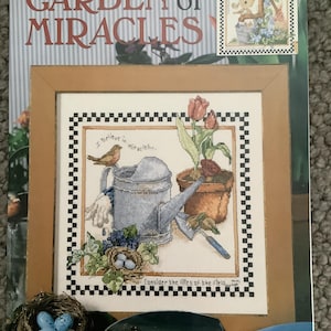 Può includere: Un libro di schemi per punto croce intitolato "Garden of Miracles" con un disegno a punto croce incorniciato con un annaffiatoio, un uccello e fiori. La copertina presenta il testo "Garden of Miracles" e "Cross Stitch".