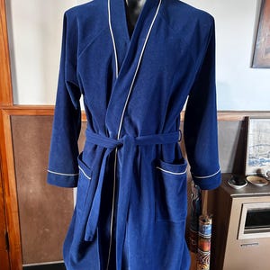May include: A deep navy blue robe featuring long sleeves, a shawl collar, and a matching tie belt at the waist. The garment is accented with thin white piping along the collar, the front opening, the sleeve cuffs, and the two front patch pockets.