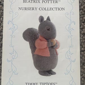 Puede incluir: Portada de libro con una ardilla tejida de la colección Beatrix Potter Nursery. La ardilla es gris con una chaqueta rosa y sostiene un objeto pequeño. El texto incluye "Beatrix Potter Nursery Collection" y "Timmy Tiptoes".