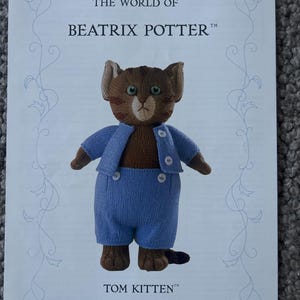 Puede incluir: Una tarjeta impresa con un gatito tejido de color marrón que lleva una chaqueta azul claro y pantalones cortos a juego con botones blancos. El texto dice THE WORLD OF BEATRIX POTTER y TOM KITTEN, enmarcado por adornos azul claro.