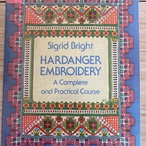 May include: A book titled "Sigrid Bright Hardanger Embroidery: A Complete and Practical Course." The cover features a blue background with a decorative border in pink, orange, green, and red. The book is part of the Dover Needlework Series.