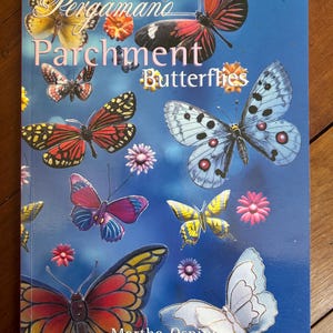 May include: A book titled Pergamano Parchment Butterflies by Martha Ospina with a blue cover featuring various colorful butterfly illustrations, including red, yellow, blue, and white species, alongside small pink and orange floral accents.