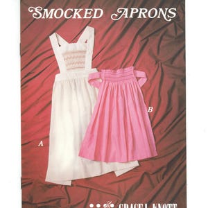 May include: Two smocked aprons, one white with a smocked design and one pink with a gathered ruffle. The aprons are on a red fabric background. The image includes the text "SMOCKED APRONS" and "GRACE L. KNOTT SMOCKING SUPPLIES LTD."