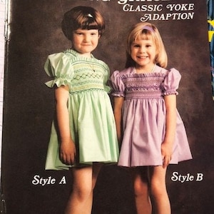 May include: Two girls wearing smocked dresses. The girl on the left is wearing a light green dress with a white smocked yoke and the girl on the right is wearing a light purple dress with a white smocked yoke. The text "Amazing Grace Classic Yoke Adaption" is at the top of the image. The text "Style A" is below the girl on the left and the text "Style B" is below the girl on the right.