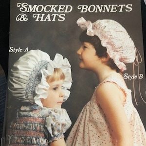 May include: Two girls wearing smocked bonnets and dresses. The girl on the left is wearing a white bonnet with a blue trim and a floral print dress. The girl on the right is wearing a pink bonnet with a floral print dress. The image includes the text "Smocked Bonnets & Hats" and "Style A" and "Style B".