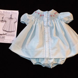 May include: A light blue baby bishop dress with pink embroidered flowers and a white button closure. The dress is made of a soft, lightweight fabric and has a gathered skirt. The dress is accompanied by a pattern for making the dress.