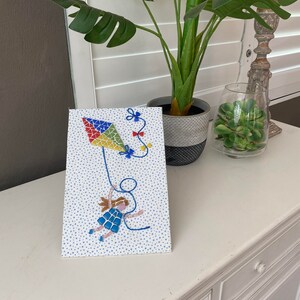 May include: A framed artwork featuring a colorful kite and a girl. The kite is a patchwork of red, yellow, green, and blue triangles. The girl is wearing a blue dress and holding the kite string. The background is white with blue stars.
