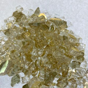 May include: A pile of small, clear glass pieces with a golden tint. The glass pieces are irregular in shape and size, and they are scattered on a white surface.