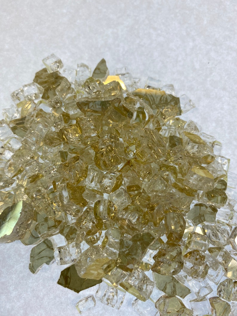 May include: A pile of small, clear glass pieces with a golden tint. The glass pieces are irregular in shape and size, and they are scattered on a white surface.