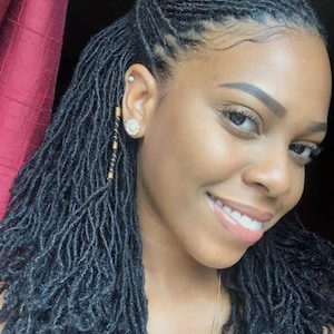 May include: A smiling person with dark skin and long, dark micro-locs. The hair is styled with some locs pulled back. The person is wearing a small stud earring and a gold earring with a dangling design. A gold necklace is also visible.