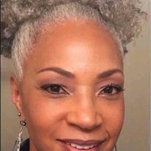 May include: A woman with short, gray, curly hair styled in a high bun. She is wearing a blue and white patterned shirt and silver earrings.