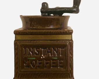 Könnte beinhalten: Ein brauner Keramikbehälter mit quadratischem Boden und passendem Deckel, der einen dunkelbraunen Kurbelgriff aufweist. Die Vorderseite des Behälters ist mit dem Text INSTANT COFFEE in Großbuchstaben geprägt und mit kleinen grün-weißen Details.