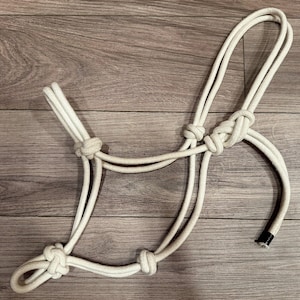 May include: A white rope halter constructed from thick, braided cord. The design features multiple fixed knots at key connection points to form a structured headpiece. One end of the rope is finished with a black plastic cap for durability.