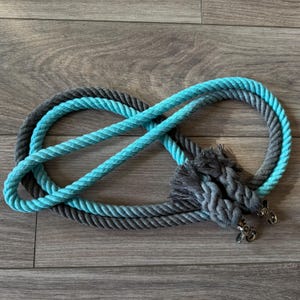 May include: A two-toned rope leash in gray and turquoise blue, looped in an infinity shape. The leash has a braided tassel at the end and silver-colored metal clasps. The rope is thick and appears to be made of cotton.