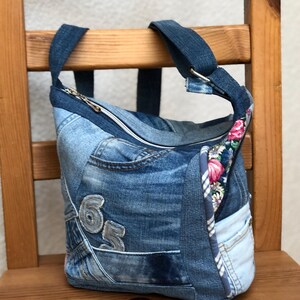 Può includere: Borsa a tracolla in denim con design patchwork. La borsa presenta una chiusura con cerniera, un accento in tessuto floreale e il numero "65" cucito sul davanti. La borsa ha una tracolla in denim ed è su una sedia di legno.