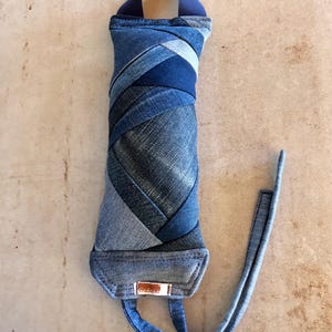 May include: A denim-covered bottle carrier with a blue handle and strap. The carrier is made from various shades of blue denim, stitched together in a diagonal pattern. A small brown tag is attached to the bottom.