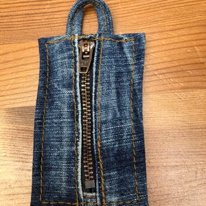 Könnte beinhalten: Kleine Denim-Tasche mit Reißverschluss und Henkel. Die Tasche besteht aus blauem Denim-Stoff mit sichtbaren Nähten in kontrastierendem gelb-braunem Faden. Unten ist ein kleines Etikett angebracht.