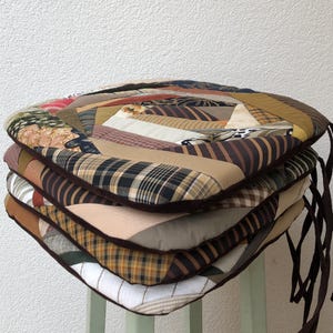 Könnte beinhalten: Ein Stapel von vier braunen, beige und weißen Patchwork-Stuhlkissen mit braunen Bändern. Die Kissen bestehen aus verschiedenen Stoffen und Mustern, darunter Karo, Streifen und Blumenmuster.