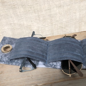 Może przedstawiać: Szary materiałowy futerał na okulary z metalowym oczkiem. Wewnątrz futerału znajdują się dwie pary okularów. Jedna para jest czarna, a druga brązowa.