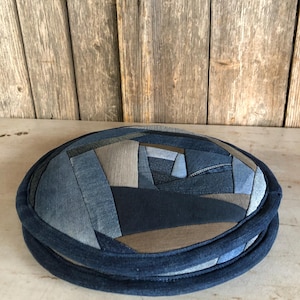 May include: A round, blue denim patchwork cushion with a dark blue border. The cushion is made from recycled denim fabric and is perfect for adding a touch of rustic charm to any room.