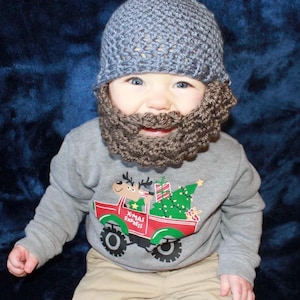 May include: A baby wearing a gray crocheted hat and a brown crocheted beard. The baby is wearing a gray sweatshirt with a red truck and a Christmas tree on it. The baby is sitting on a blue blanket.