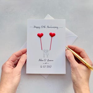 May include: A white anniversary card with the text "Happy 12th Anniversary". The card features two red heart-shaped balloons held by stick figures. The names "Adam & Lauren" and the date "14-07-2012" are also printed on the card.