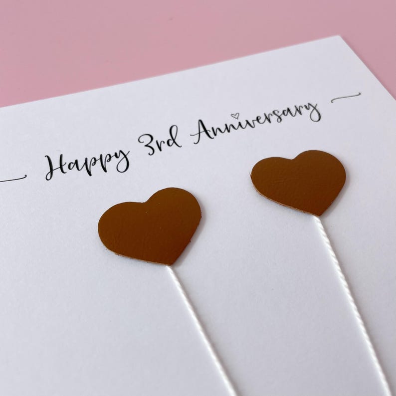 May include: A white greeting card with the text "Happy 3rd Anniversary" in black script. Two bronze-colored heart-shaped embellishments are attached with white strings. The background is a soft pink.