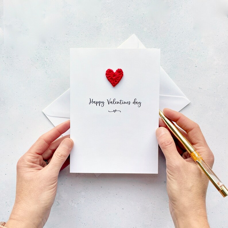 A white card with 'Happy Valentines Day' printed across the middle. This is printed in black cursive text. Above the text is a cute red crochet heart.