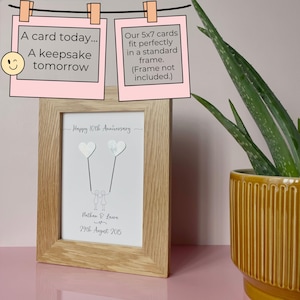 Op de afbeelding: Een ingelijste print met een licht houten frame. De print toont een tekening van twee figuren die hartvormige ballonnen vasthouden, met de tekst "Happy 10th Anniversary", namen en een datum. Bovenin staan twee roze kaarten met "A card today... A keepsake tomorrow" en "Our 5x7 cards fit perfectly in a standard frame."