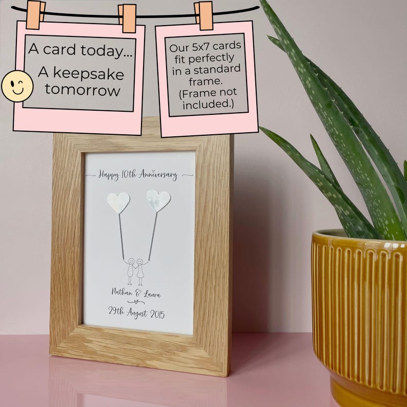Op de afbeelding: Een ingelijste print met een licht houten frame. De print toont een tekening van twee figuren die hartvormige ballonnen vasthouden, met de tekst "Happy 10th Anniversary", namen en een datum. Bovenin staan twee roze kaarten met "A card today... A keepsake tomorrow" en "Our 5x7 cards fit perfectly in a standard frame."