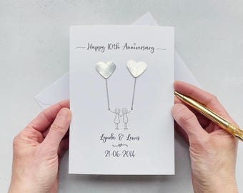 May include: A white anniversary greeting card featuring the text Happy 10th Anniversary, two silver heart-shaped embellishments attached to thin lines held by two stick figures, the names Lynda & Lewis, and the date 21-06-2014 printed in black ink.