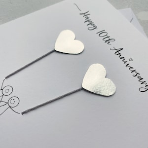 Op de afbeelding: Een witte jubileumkaart met de tekst "Happy 10th Anniversary" in zwart script. Twee zilveren hartvormige decoraties zijn bevestigd met grijze draad. Stippelfiguren zijn onderaan de kaart getekend.