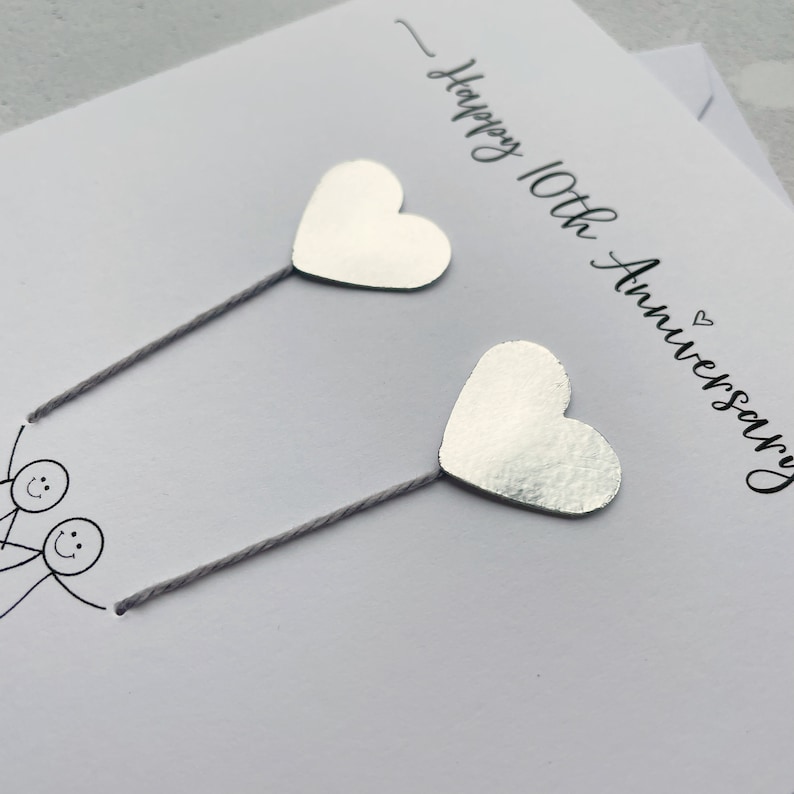 Op de afbeelding: Een witte jubileumkaart met de tekst "Happy 10th Anniversary" in zwart script. Twee zilveren hartvormige decoraties zijn bevestigd met grijze draad. Stippelfiguren zijn onderaan de kaart getekend.