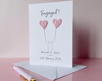 以下が含まれることがあります： 上部に「Engaged!」と書かれた白いグリーティングカード。その下には、2つの薄いピンクのかぎ針編みのハートにつながった紐を持つ2人の棒人間が描かれています。カードにはHannah & Jamesという名前と2026年2月14日という日付が黒いインクで記されています。